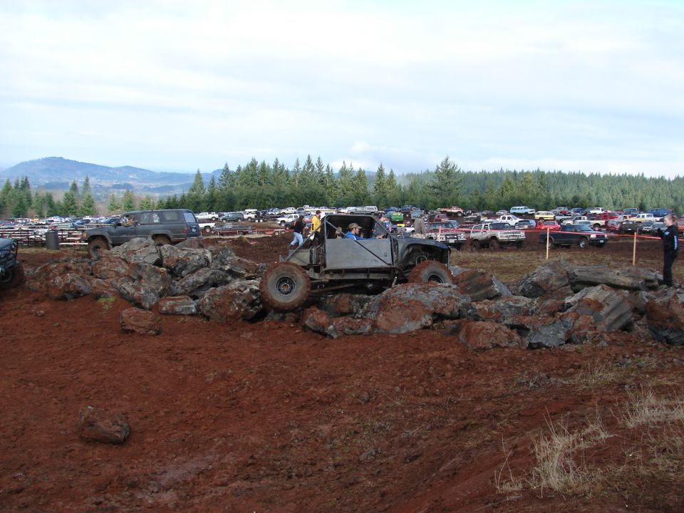Custom off-road buggy with roll cage and large tires climbing over rocks, with many other vehicles parked in the background