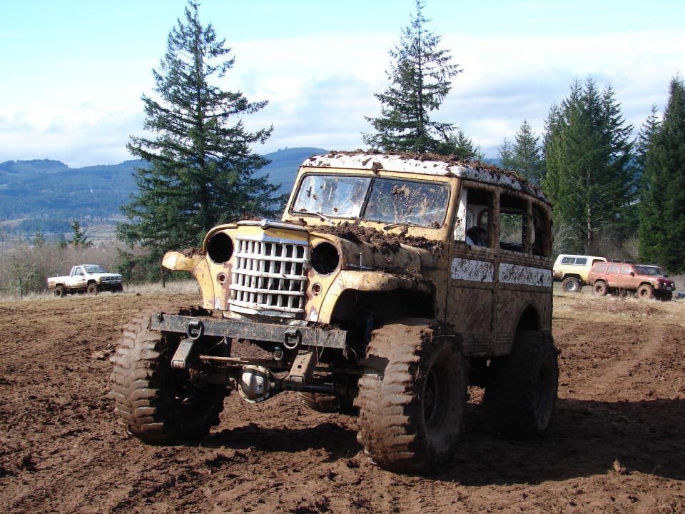 Mud-covered vintage Jeep wagon with large tires in a muddy field; white pickup truck and two SUVs in background