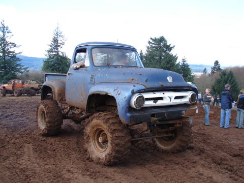 1996 Ford Bronco - A lifted 1956 Ford F-100 truck navigates a muddy course, with other vehicles and spectators in the background.