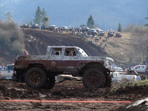 1996 Ford Bronco - A heavily modified pickup truck navigates a muddy track during an off-road event with spectators on a hill in the background.