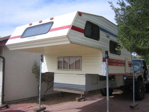 1999 Ford F-250 - A camper shell is mounted on a truck, supported by jacks, with a tree to the right.