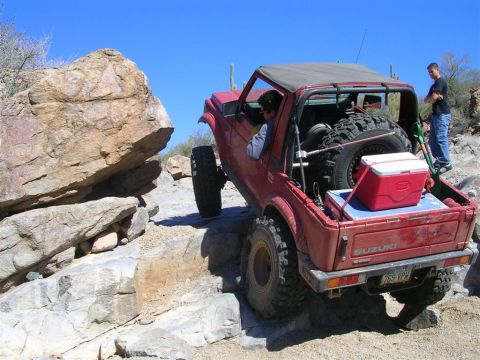 1987 Suzuki Samurai - A red Suzuki Samurai tackles a challenging rocky terrain with a driver and passenger observing.