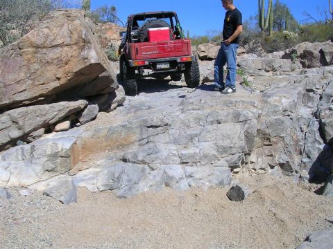 1987 Suzuki Samurai - A red 1987 Suzuki Samurai navigates a challenging rocky trail with a man standing nearby.