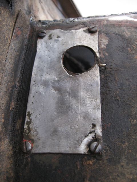 1986 Ford Bronco - A close-up view of a metal identification plate with a circular hole and visible stamped text, attached to a dark, weathered surface.