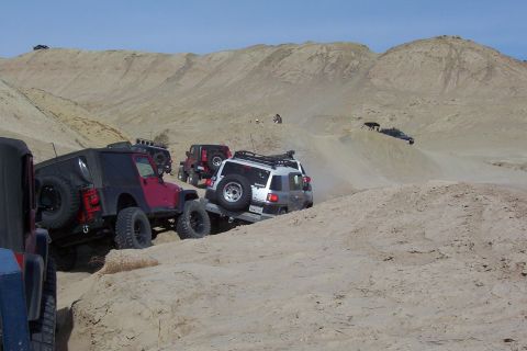 1995 Ford Bronco - Multiple off-road vehicles traverse a desert landscape, kicking up dust on the sandy hills.
