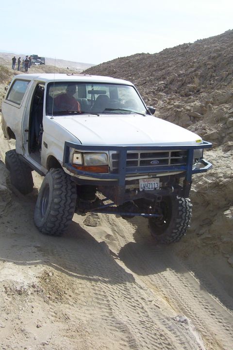 1995 Ford Bronco - A white Ford Bronco navigates a sandy terrain, with other vehicles and people visible in the background.