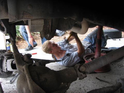 1974 Ford F-100 - A person works on the underside of a vehicle, with another person visible in the background.