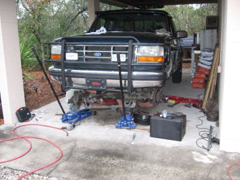 1974 Ford F-100 - Dark green Ford truck sits nose-high in a carport with the front end disassembled, front wheels and hubs pulled off to expose the brake rot…