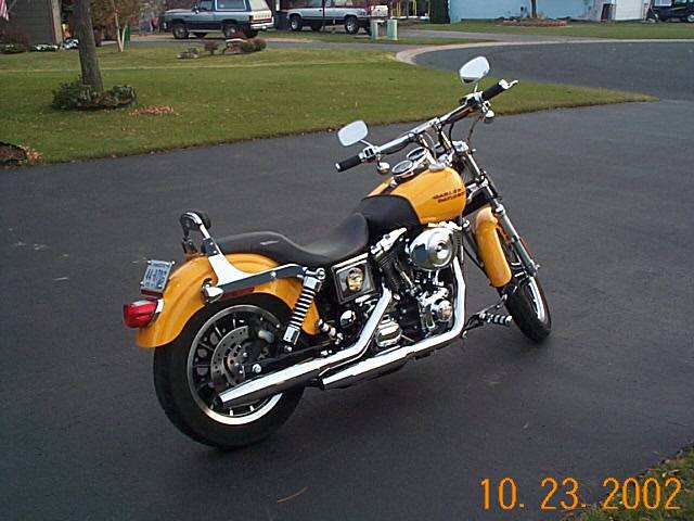 Yellow motorcycle parked on asphalt, rear three-quarter view. Chrome exhaust, black seat, license plate visible
