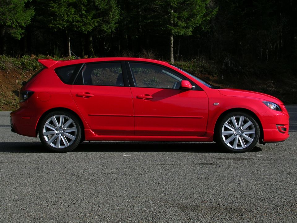Red Mazda 3 five-door hatchback in profile on asphalt with forest behind
