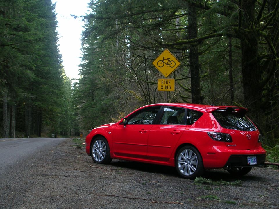 Red Mazda 3 hatchback rear three-quarter parked by forest road under a Bike Xing sign