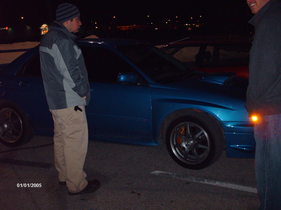Blue Subaru WRX STI with orange brake calipers and a hood scoop, parked at night with two people nearby
