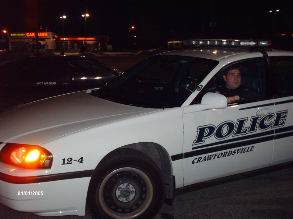 White police car with "POLICE" and "CRAWFORDSVILLE" text, parked next to another car