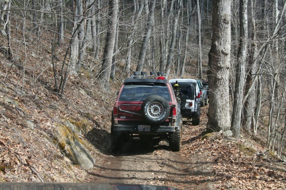 Line of off-road SUVs driving on a wooded trail. The lead vehicle is a red SUV with a spare tire mounted on the back