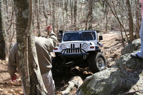 1979 Ford Bronco - A white Jeep Wrangler is shown off-roading on a challenging, rocky path through a wooded area.