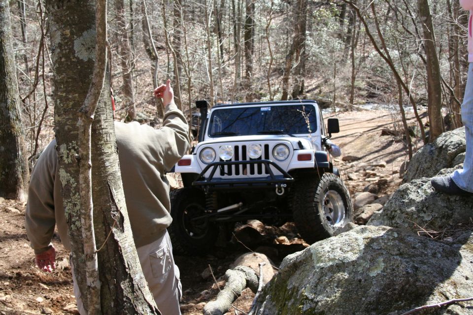 White Jeep Wrangler navigating a rocky, wooded trail. A person points ahead