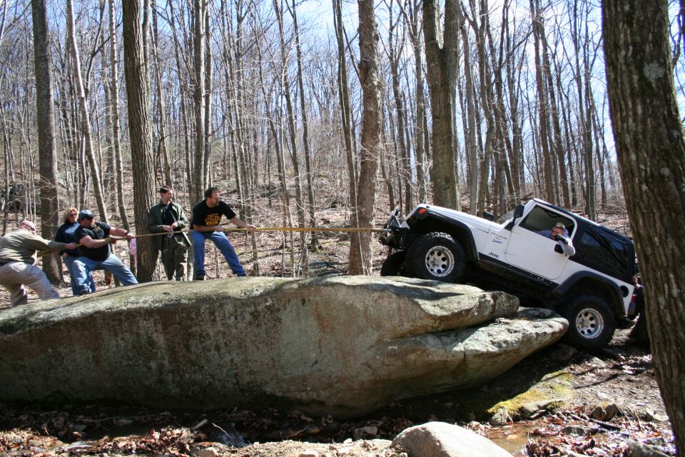 White Jeep Wrangler hung up on a large rock in the woods while several people pull it with a tow strap