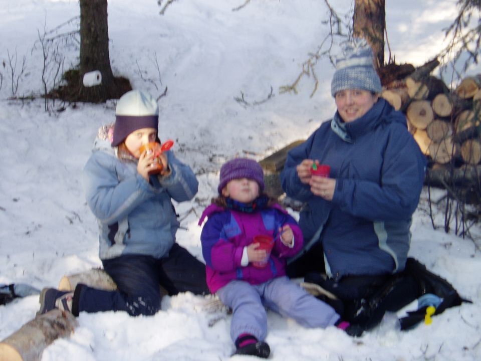 Three people in winter clothing sitting in the snow, two children drinking from cups and an adult holding a cup