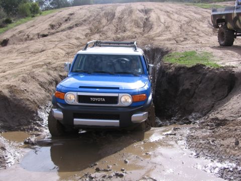 1989 Ford Bronco - A blue Toyota FJ Cruiser navigates a challenging muddy off-road course, with another vehicle in the background.