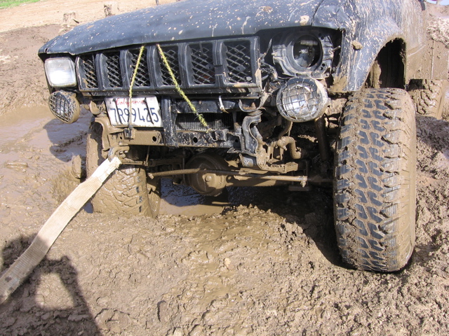 Front of a dark-colored SUV stuck in deep mud, with a tow strap attached