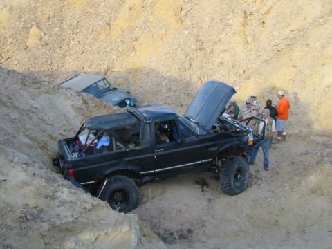 1992 Ford Bronco - Two off-road vehicles are stuck in a steep dirt embankment, one with its hood open.