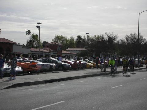 1998 Ford Crown Victoria - row of sports cars parked at a lot car show while several cyclists ride past on the adjacent street.