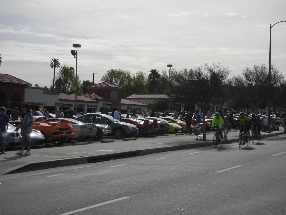 Cars lined up at a parking-lot car show with cyclists riding past on the street