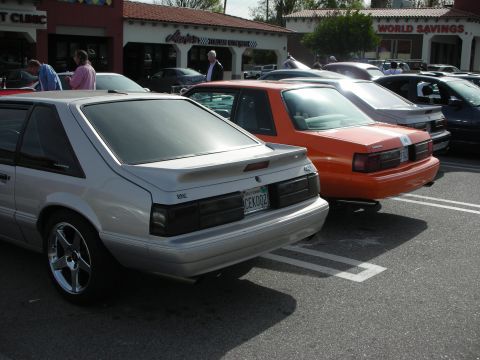 1998 Ford Crown Victoria - silver and an orange Fox-body Ford Mustang hatchback parked at a strip-mall car show near a 'World Savings' storefront.