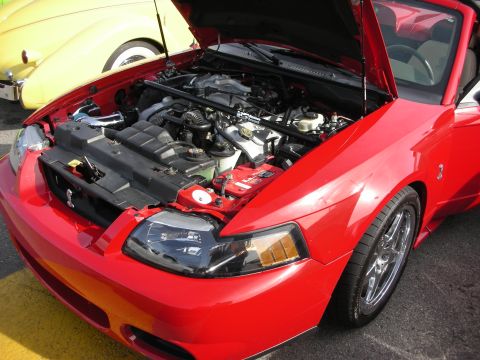 1998 Ford Crown Victoria - open engine bay of a red Ford Mustang showing a V8 with a cold-air intake and chrome wheels, at an outdoor show.