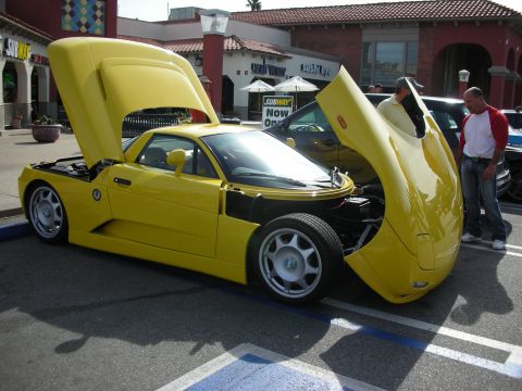 1998 Ford Crown Victoria - yellow mid-engine sports car with its scissor doors and rear hatch open, parked outside a strip mall.