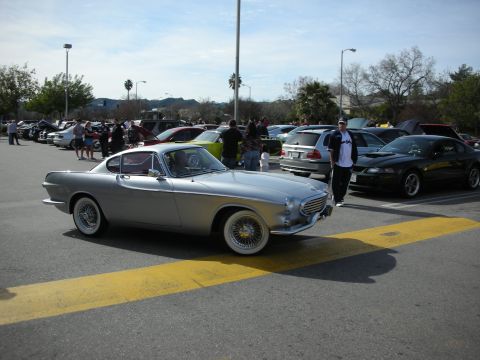 1998 Ford Crown Victoria - silver Volvo P1800 coupe with wire wheels rolling through a crowded outdoor car show parking lot.