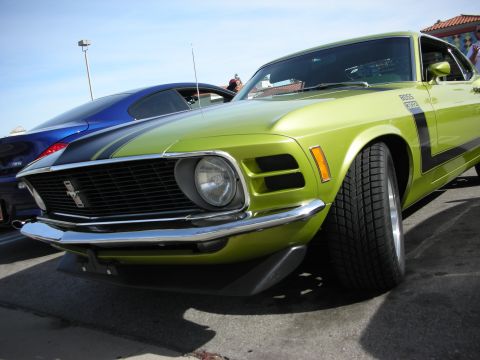 1998 Ford Crown Victoria - lime-green Boss 302 Mustang fastback parked beside a blue muscle car at a sunny outdoor car show, showing its quad-headlight grille.