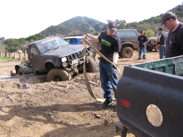 guy pulling tow strap from stuck off-road truck in mud, other trucks and people around