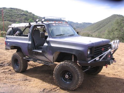 1989 Ford Bronco - Modified SUV with off-road tires and a roof rack sits on a dirt track with hills in the background.