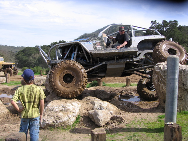 Modified off-road buggy with large tires navigating a rocky obstacle course; another yellow off-road vehicle is visible in the background