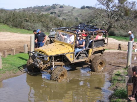 1989 Ford Bronco - A yellow off-road vehicle navigates a muddy terrain with several passengers.