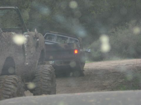 1989 Ford Bronco - Rain-spotted windshield shot of two mud-caked lifted 4x4s on a wet dirt trail, the nearer one a mud-covered rig on huge knobby tires. The d…