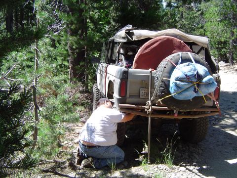 1967 Ford Bronco - A person kneels to work on the rear bumper of a modified Ford Bronco parked on a dirt road in a wooded area.