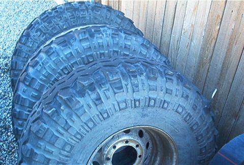 1986 Ford Bronco - Four off-road tires are stacked and leaning against a wooden fence.