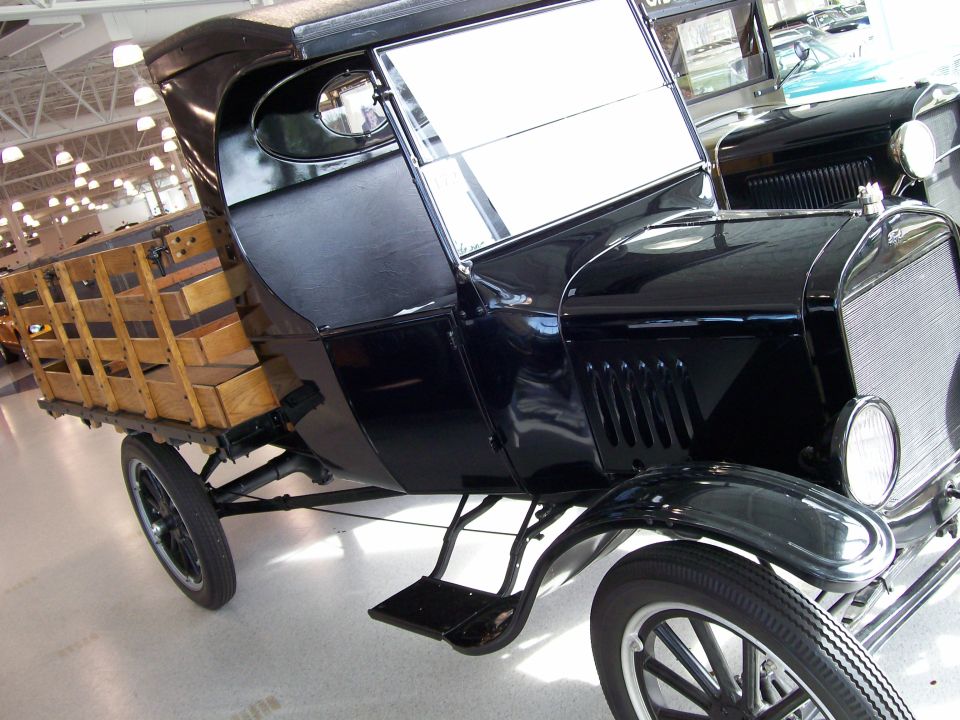 Black antique truck with a wooden bed filled with boxes. Another vehicle is partially visible behind it