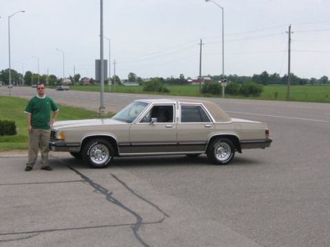1988 Mercury Grand Marquis - Full side profile of a tan/champagne Mercury Grand Marquis four-door sedan parked in a lot beside a rural highway, with a man in a green po…