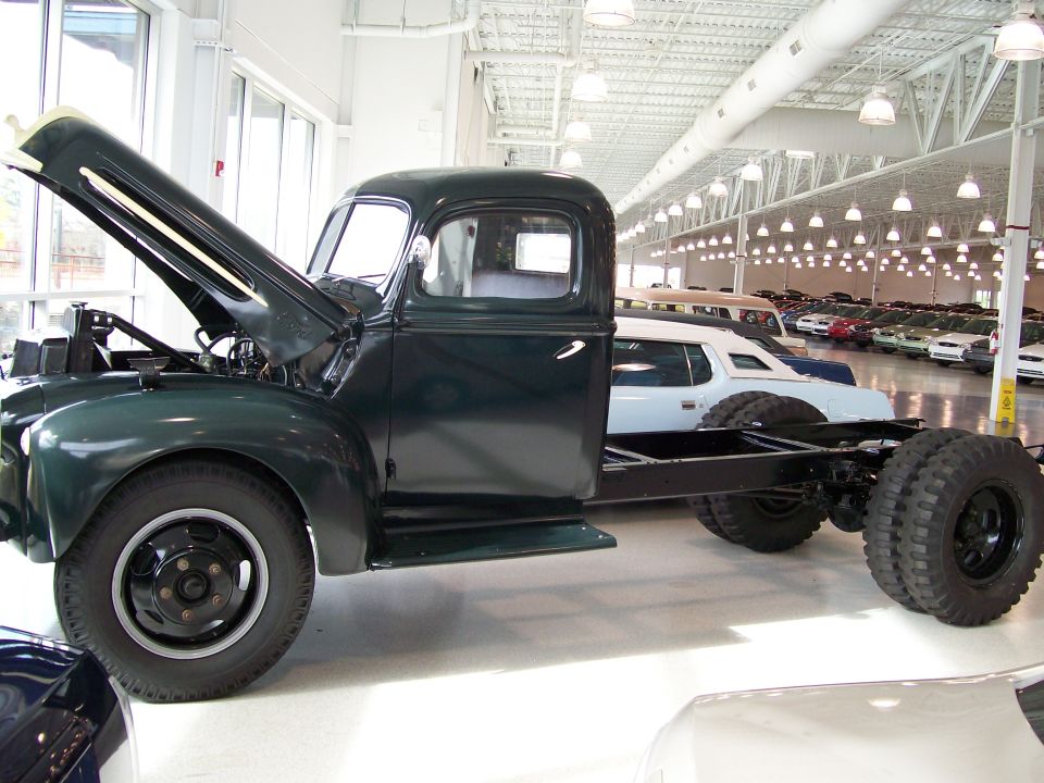 Dark green truck chassis, hood open, side view. Dual rear wheels visible