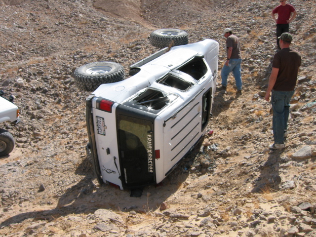 Overturned white SUV with large tires on a rocky desert trail. Two other vehicles and three people are visible