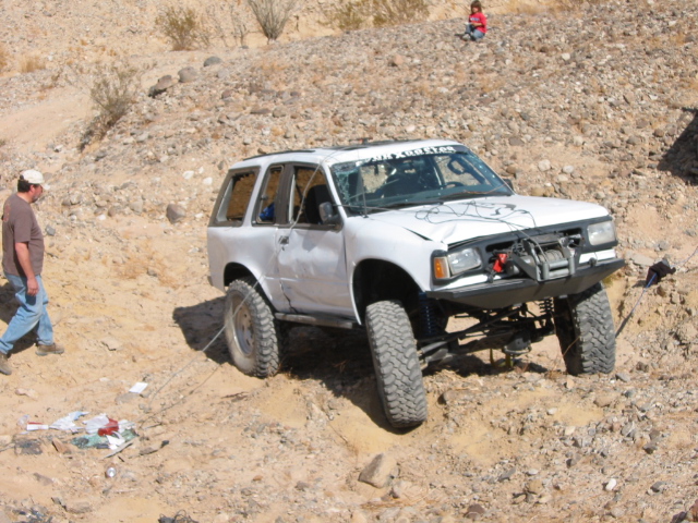 White off-road SUV with a winch and aftermarket bumper, damaged windshield, and lifted suspension, stuck on a dirt hill