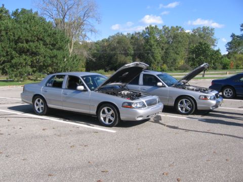 1988 Mercury Grand Marquis - Two silver Mercury Grand Marquis sedans are parked side-by-side with their hoods open, revealing their engines.