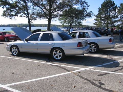 1988 Mercury Grand Marquis - Two silver sedans are parked in a lot near a body of water, with one having its hood up.