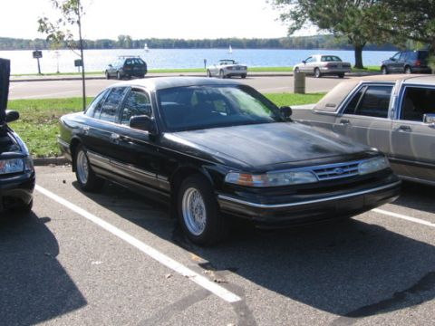 1988 Mercury Grand Marquis - A black sedan is parked in a lot next to a body of water, with other vehicles visible in the background.