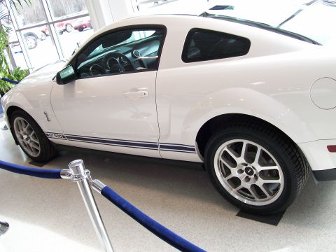1996 Ford Bronco - A white Ford Mustang GT500 is parked indoors, possibly in a showroom, with blue velvet ropes in the foreground.