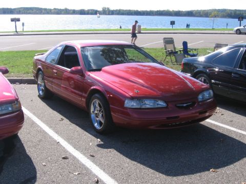 1988 Mercury Grand Marquis - A red coupe with a large hood scoop is parked next to another car near a body of water.