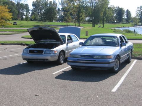 1988 Mercury Grand Marquis - A blue 1988 Mercury Grand Marquis is parked in a lot next to a silver sedan, whose hood is open.